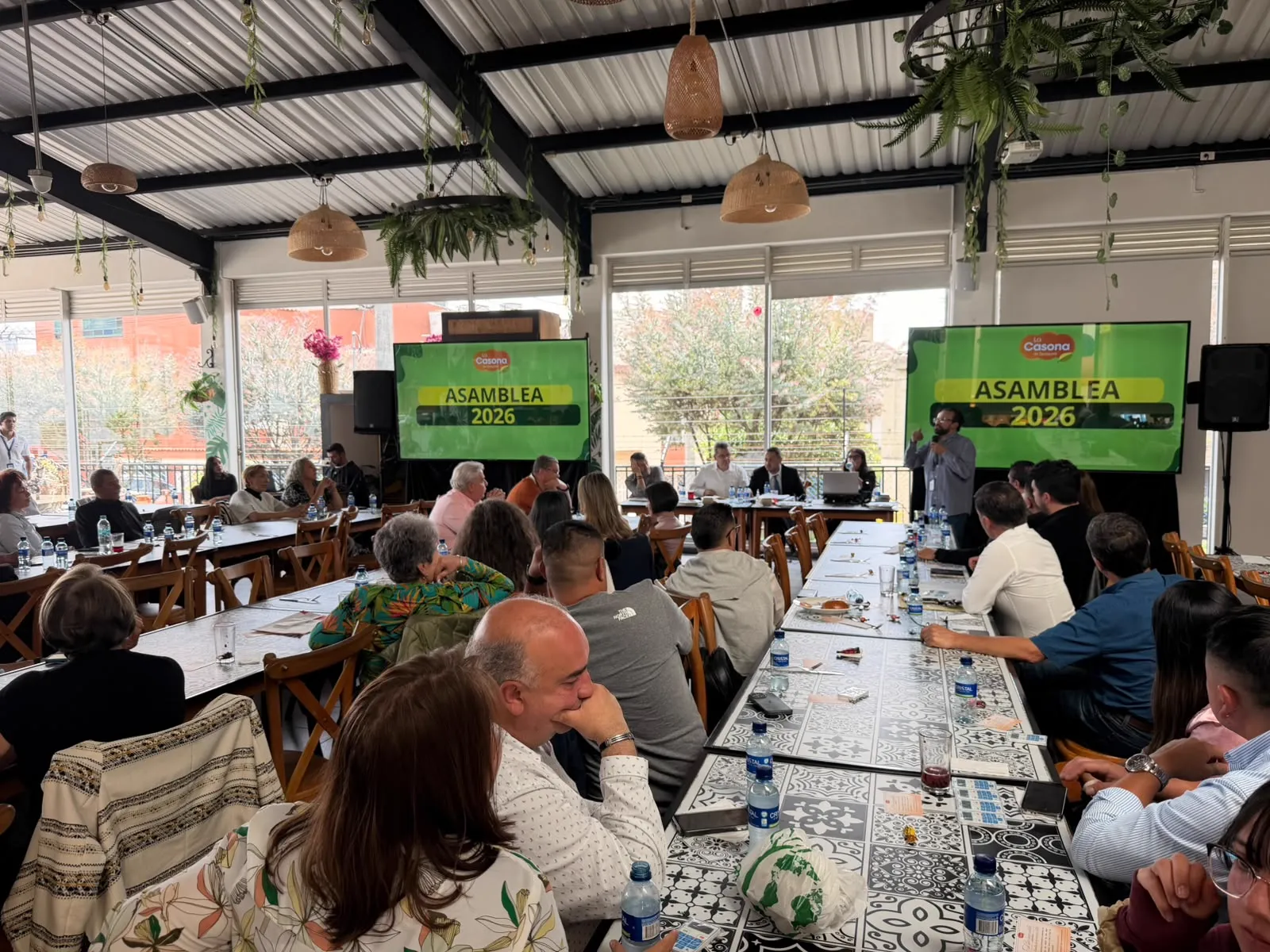 Asamblea 2026 de La Casona operada por CLICK: asistentes en mesas largas, mesa directiva al fondo frente a los ventanales, dos pantallas con la marca de la empresa y el texto «Asamblea 2026», parlantes en trípode y un orador hablando con micrófono