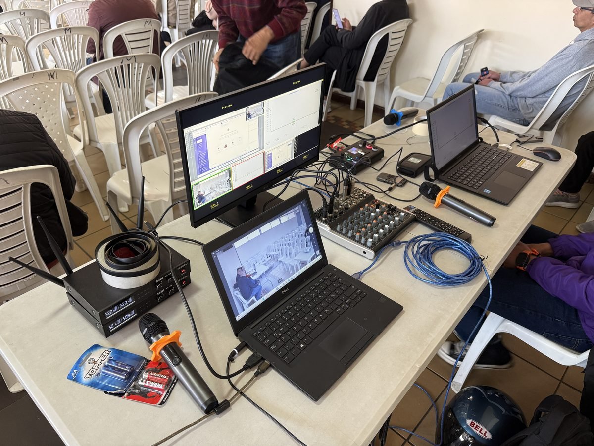 Mesa técnica de CLICK en un salón comunal antes de una asamblea de copropietarios: monitor con las cámaras y la presentación, consola de sonido, receptores de micrófonos inalámbricos, dos micrófonos de mano y las sillas tipo auditorio al fondo