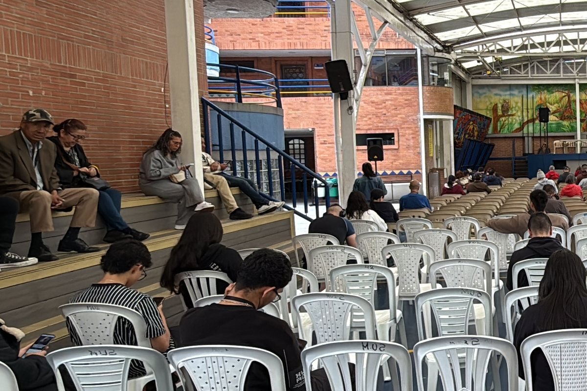 Salón de una asamblea de copropietarios operada por CLICK, visto desde atrás: propietarios sentados en filas de sillas blancas y en las gradas de la izquierda, esperando el inicio; al fondo, el escenario con un parlante