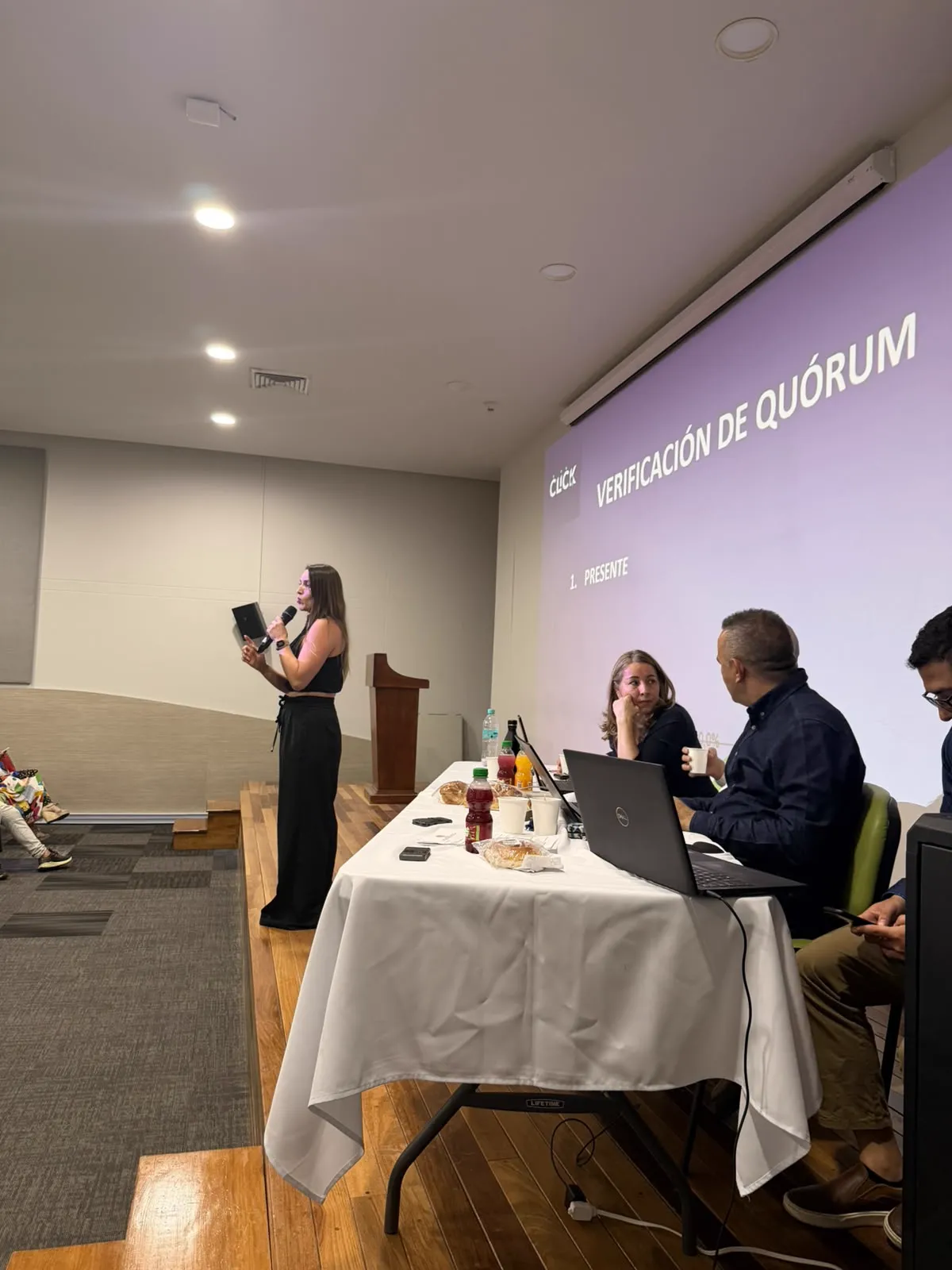 Mujer hablando con micrófono junto a la mesa directiva, con la verificación de quórum proyectada en pantalla, en una asamblea operada por CLICK